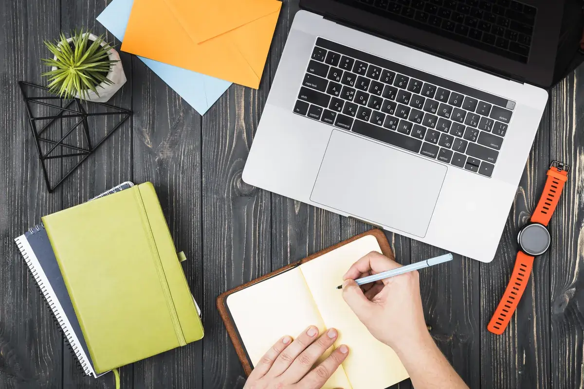A desk with a laptop, notebooks and a person writing