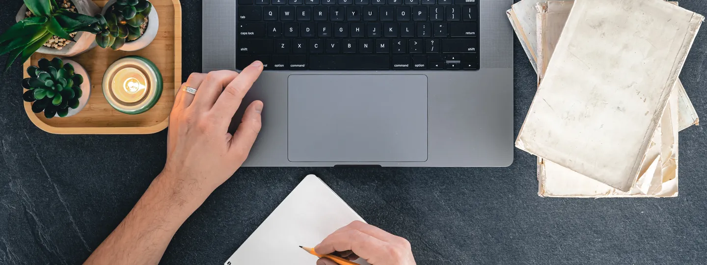 Vue de dessus d'un bureau sombre : une main sur un ordinateur portable, l'autre prend des notes